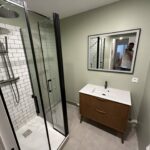 Salle de bain rénovée avec cabine de douche en verre, meuble-lavabo en bois, et miroir par Al'eau Nico.