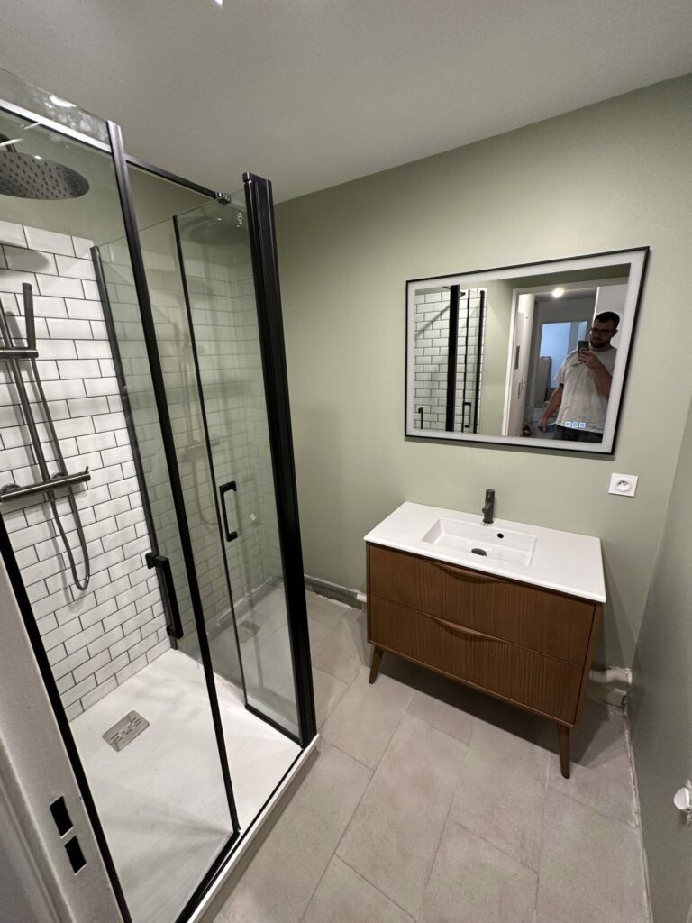 Salle de bain rénovée avec cabine de douche en verre, meuble-lavabo en bois, et miroir par Al'eau Nico.