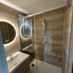 Rénovation moderne et élégante d'une salle de bain avec douche à l'italienne transparente, meuble vasque suspendu en bois, et miroir LED circulaire par Al'eau Nico et Eau Secours.